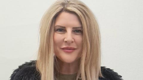 Councillor Maxine Clark - a woman with long blonde hair and wearing a black feathery top with a green shirt - smiling as she stands in front of a white wall.