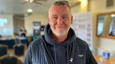 Chris Jones. Man in his 50s with facial hair, in a dark navy hooded jumper and coat, and wearing a Prostate Cymru charity badge.