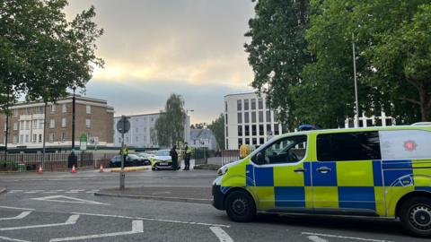A police van is parked on a road opposite the entrance to a car park, where a police car is parked. Two officers can be seen at the entrance, with one speaking to someone dressed in black. There are trees and buildings surrounding the car park, which has a few civilian cars parked in it.