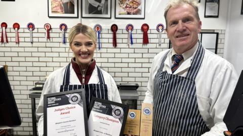 A woman with tied-back blonde hair and a man with short fair hair are both dressed in wite coats and blue-and-white striped butcher's aprons. They are in a white-tiled butchery with rosettes along the top of the tiles and framed product photos above. The pair are holding framed certificates and wooden awards in their hands
