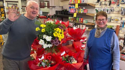 A man with short hair and wearing a grey jumper and a woman with short hair and dark framed glasses in a blue jumper and blue pinafore are smiling and at the camera next to large bunches of Valentine's day flowers