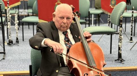 George Latham wearing a black blazer with a white shirt, black braces and grey trousers. He is also wearing a navy blue tie with diagonal red stripes. He is playing a cello while looking at the camera.