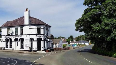 The Ginger Hall pub, a large white building with a section of the TT course running past it. There are tree on the other side of the road.