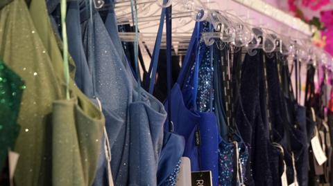 A colorful array of glamorous and sparkly prom dresses hung up in a shop they each have a tag on.