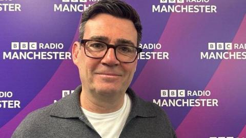 Andy Burnham, 56, who has black hair with streaks of grey and wears black framed galesses, stands in front of a purple screen bearing the BBC Radio Manchster logo. He is wearing a grey button up jumper over a white t-shirt. 