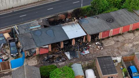 A drone image shows a row of garages, with burned and charred debris either side of the building. One of the garages has a corrugated iron roof, where the others have felt roofs. None has been destroyed by the fire. A concrete retaining wall, with the A47 below, can be seen in the upper right of the image. Other garden buildings, garages and vehicles can be seen in the foreground and lower left of the image.