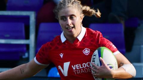 Maddy Grant in action for Canada, sporting a braided light-haired ponytail, wearing a red shirt, cradling a rugby ball with her left hand and arm
