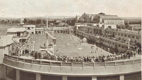 A black-and-white photo shows Morecambe Super Swimming Stadium, with crowds sitting in the grandstands around a large pool. 