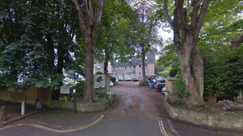 A Google Maps picture of the hotel, a three-storey building seen from a road. Two large trees sit either side of the entrance road