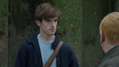 Jude stands in front of a mossy wall, facing another character whose face we can't see. It is Detective Inspector Max Arnold, played by Adrian Scarborough, who has ginger hair and wears a green coat. Jude wears a blue t-shirt and navy collared jacket, with a brown satchel strap across his body. He had brown hair swept over to one side. 