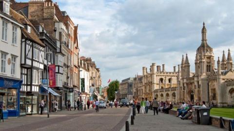King's Parade in Cambridge. To the left is a row of shops and to the right is the frontage of King's College. People are walking, cycling and driving on the road in between the two. 