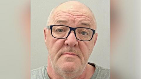 Police custody image of David Berry with receding grey hair and grey beard wearing black glasses and grey t-shirt. 