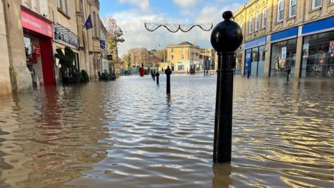 Low down view of water in Chippenham town centre, bollards poking up from it and the bottom of shops clearly engulfed.