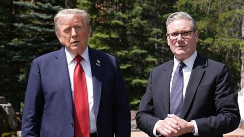 Donald Trump has white hair and is wearing a dark blue suit jacket, white shirt and red tie. He is standing in front of trees next to Keir Starmer who has grey hair and is wearing a black suit jacket, white shirt, grey tie. He is wearing glasses.