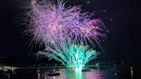 Fireworks have been ignited over Plymouth Sound. There are emerald green sprays of light lower to the ground and a mass of violet fireworks have exploded above them. There are some studs of bright blue falling making a downward curve. The colours are reflected in the water and illuminate boats full of spectators.