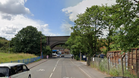 A long road with an arched railway bridge. There is a green space on the left and metal containers behind a metal fence on the right, as well as green trees and bushes