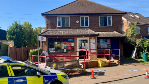A residential street with a police presence outside a two-story brick building, used as the post office. A police car is parked at the roadside, surrounded by traffic cones that cordon off part of the area. The sky is partly cloudy, casting soft light over the scene.