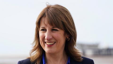 Rachel Reeves on a visit to Southport Pier - her background is obscured but it is clear she is standing outside - she is smiling with her top teeth visible, and is wearing a dark coloured blazer and a royal blue shirt