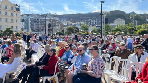A picture of people sat down on chairs. There is a number of people in hats and holding flags