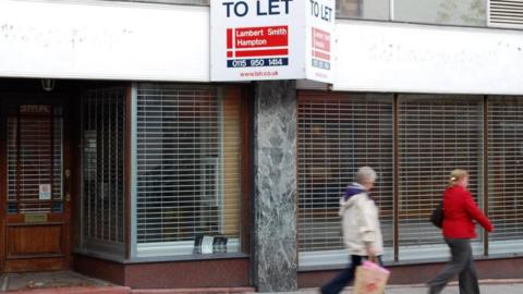 Two women walk past a shuttered shop. A 'to let' sign is on display above it.