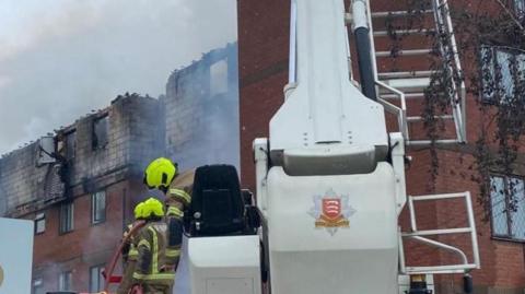 Two firefighters operate an aerial ladder platform as they tackle the fire. The four-storey block of flats is pictured in front of them. It is an orange brick building but its top floor is totally charred and damaged by the fire.