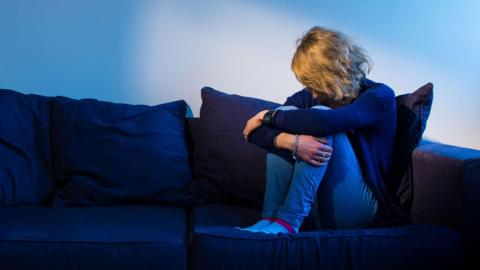 A woman sat on a sofa holding her knees with her head down. The sofa is blue and she is wearing blue jeans and a blue top. 