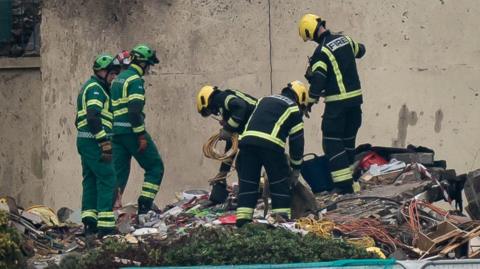 Two paramedics in green uniforms and three firefighters in black uniforms search through rubble left behind after an explosion in Jersey.