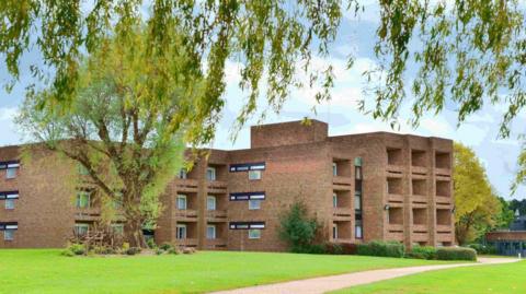 Brick building with 28 visible windows across three stories. There is a pavement passing through a grassed areas and a large tree.