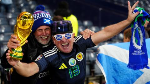 Two smiling Scotland fans during a match in Hampden Stadium, Glasgow. One is holding a replica World Cup and a scarf, and is wearing sunglasses with a saltire pattern. The other has a hat with Scotland written on it.