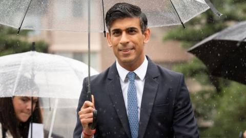 Rishi Sunak walks towards the camera during his visit to the G7 summit in Hiroshima, Japan. He's holding a clear plastic umbrella, dotted with raindrops. There are other people holding umbrellas in the background