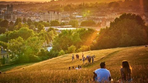 An orange evening sunset in Bath looking over hills. In the bottom right corner there is a couple sat together looking at the view, facing away from the camera