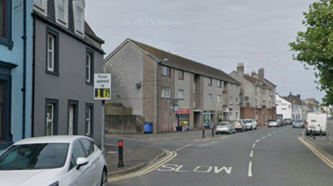 A street with grey buildings on one side and trees on the other. The road has "slow" written on it with a speed monitor on the left
