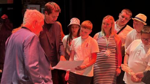 The TV presenter is standing in a crowd looking at a drawing of a young person's housing design