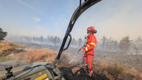 A firefighter in bright red protective clothing stands in a smoke-filled heathland, spraying water onto scorched ground after a fire. The scene is viewed from inside an off-road vehicle, with part of the vehicle frame visible in the foreground. Burnt vegetation and a line of smoke-shrouded trees stretch across the background under a pale blue sky.