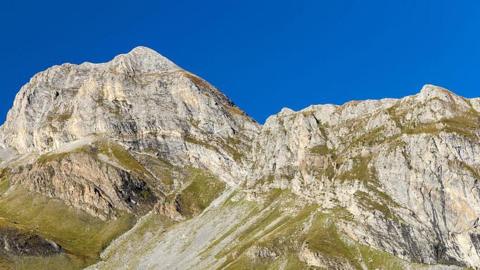 A large mountain with green vegetation at the bottom. The sky is bright blue behind it.