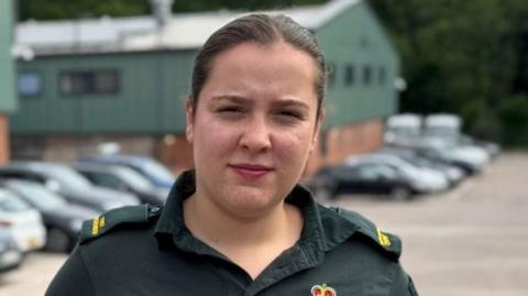 Emily Hilton wearing her dark green paramedic uniform of polo shirt, trousers and belt with badge attached. She has long dark hair tied back in a pony tail and poses with her hands behind her back next to a row of parked ambulances on one side and cars on the other