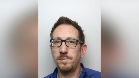 A police mugshot of David Masters, who has ginger hair and a ginger goatee. He is wearing black-rimmed glasses in the photo. His expression is neutral.