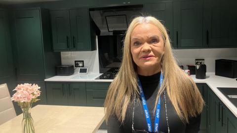 Photo of Gaynor Hutton in the kitchen of her home in Collyhurst. She has long blonde hair and wears a black long-sleeved top.