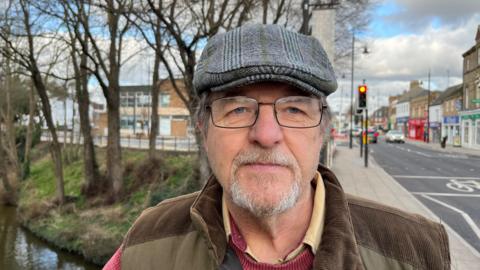 Alex Cox is 64, he has a grey thinning beard and moustache he has dark rimmed spectacles on and grey tartan/tweed flat cap on his head with grey hair sticking out slightly from the sides. He is wearing a brown body warmer over a mauve jumper with a yellow open-necked shirt underneath.