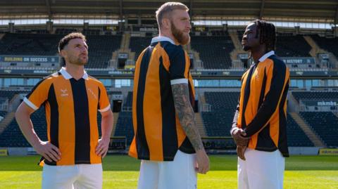 Three men stand on a football pitch wearing amber and black shirts with white shorts. They are all staring into the distance, looking in different directions. Behind them is a football stadium, with black seats in a stand.