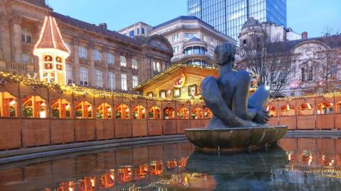A row of wooden Christmas market huts in the middle of a city centre, lit up with Christmas lights. It is viewed from the middle of a fountain with a statue right in front of us.