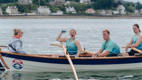 A female rower in a light blue/turquoise top punches the air while the rest of her crew look tired around her
