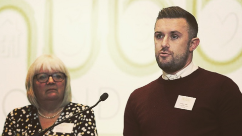 Adam Yates, wearing a burgundy jumper, speaks into a microphone as Carol Shanahan watches on.