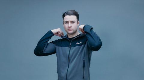 Denon Reed, who has black hair combed back, wearing a grey and black tracksuit top zipped up, standing in front of a grey backdrop. He has his hands at his collar as if adjusting it.