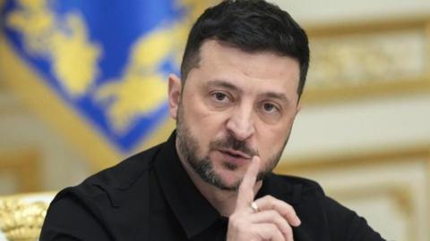 A head and shoulders image of Volodymyr Zelensky wearing his usual black suit and holding his index finger near his mouth. He has a near beard and moustache.