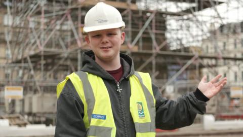 A boy in a white hard hat, a yellow hi-vis, a dark green jacket and maroon t-shirt smiles at the camera with his left hand outstretched. He is standing in front of a building site and scaffolding.