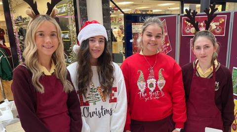 Four A-Level students are smiling at a charity stall in a shopping centre. They are wearing Christmas jumpers on top of a maroon school uniform 