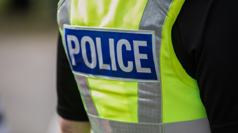 A stock image showing the back of a police officer. They are wearing a green and yellow high-visibility tabard over a black top. The word "POLICE" is emblazoned on the back.