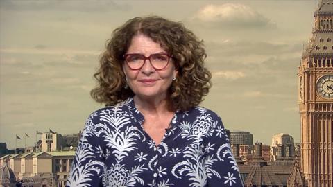 Dame Diana Johnson MP wearing red-framed glasses and a blue patterned shirt is seated in a studio setting with a backdrop of London featuring the Elizabeth Tower (Big Ben) and surrounding buildings.