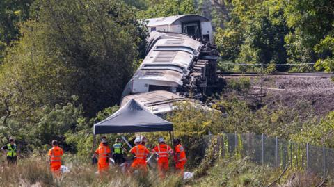 Railway carriages on their side, stretching from a railway line down the side of a small embankment. Emergency workers crowd at the bottom of the embankment.